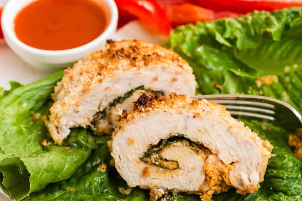 close up of a cooked and sliced buffalo chicken roll up on a bed of lettuce with buffalo sauce and veggies in the background
