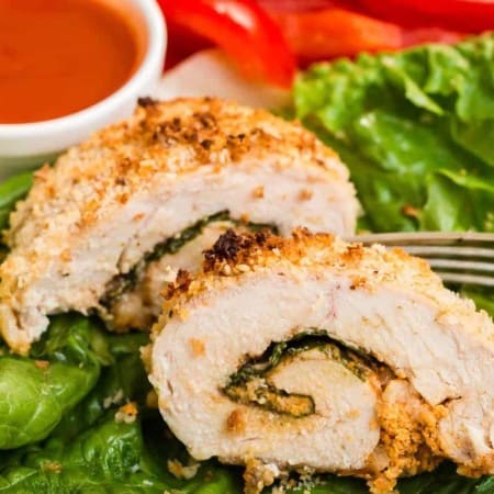 close up of a cooked and sliced buffalo chicken roll up on a bed of lettuce with buffalo sauce and veggies in the background