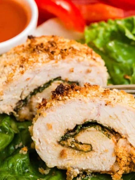 close up of a cooked and sliced buffalo chicken roll up on a bed of lettuce with buffalo sauce and veggies in the background