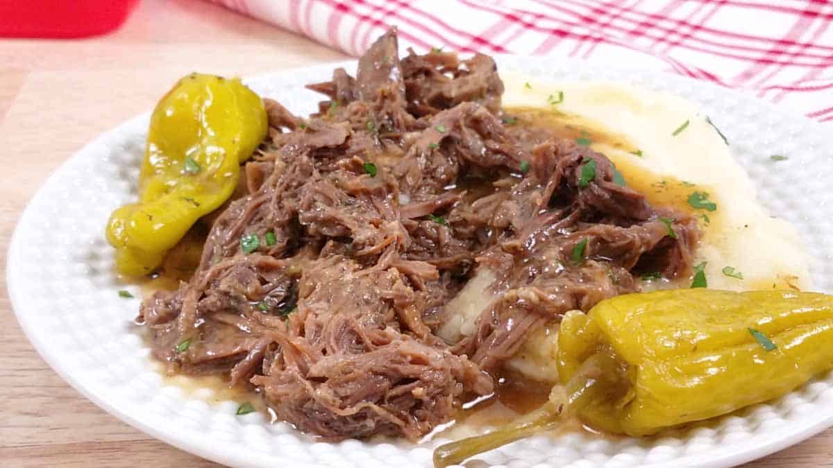 White plate with cooked Mississippi Pot Roast, pepperoncinis, mashed potatoes, and parsley sprinkled on the meat. 