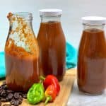 Three jars of homemade barbecue sauce on a wood cutting board with coffee beans and peppers scattered around.