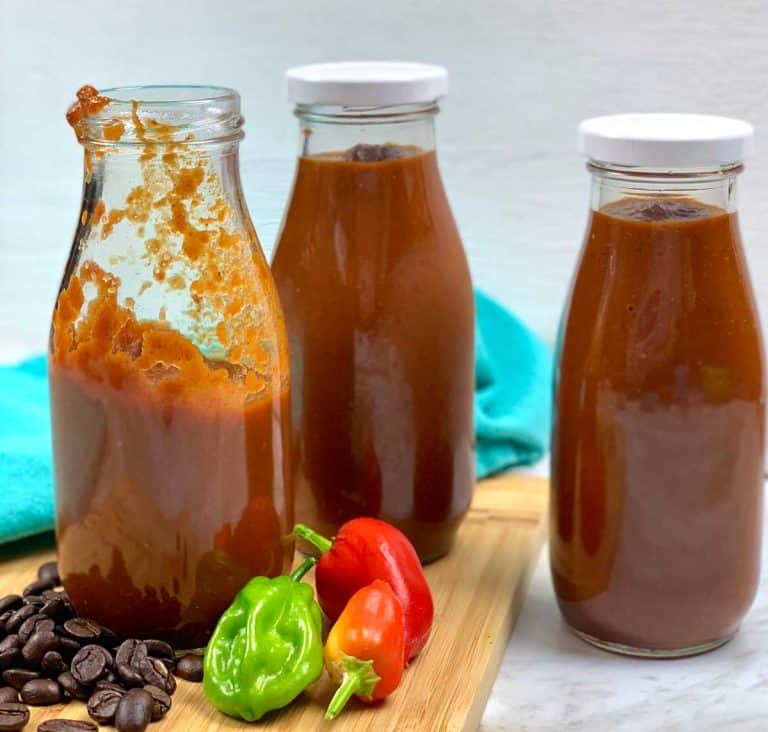 Three jars of homemade barbecue sauce on a wood cutting board with coffee beans and peppers scattered around.