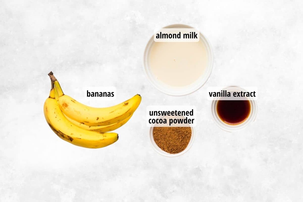 A marble counter with three bananas, and bowls of almond milk, cocoa powder, and vanilla extract.