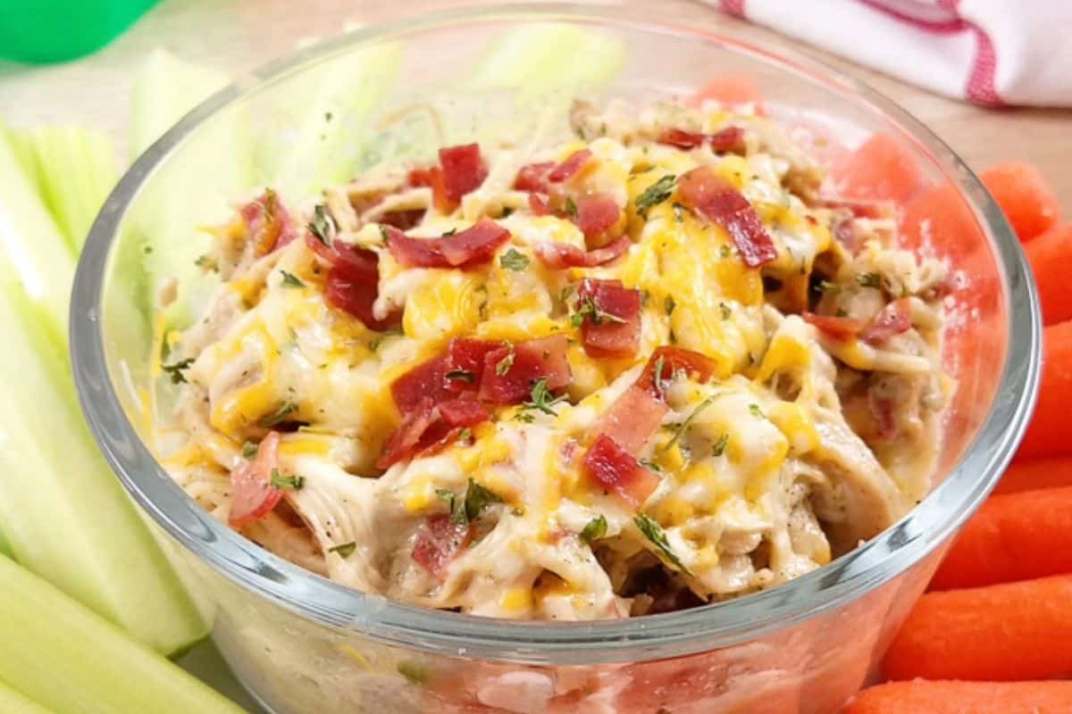 A glass bowl of cheesy, ranch chicken dip surrounded by celery sticks and baby carrots.