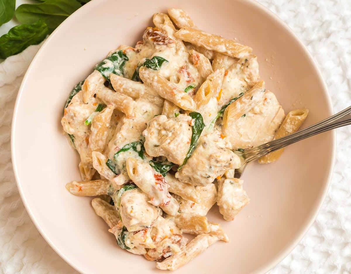 white bowl on top of a white napkin with pasta, chicken, spinach and sun dried tomatoes.