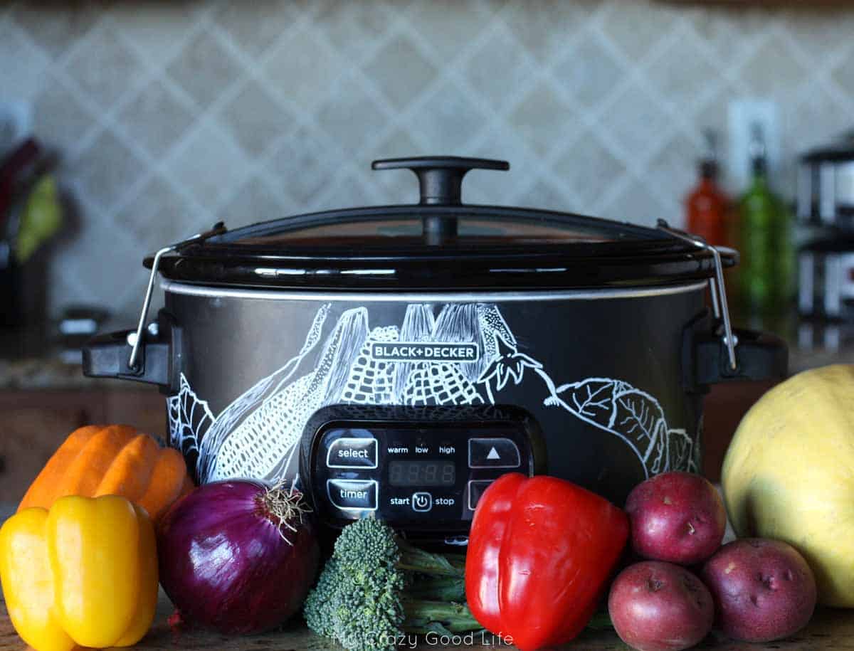 Fresh vegetables in front of a black crockpot with chalk drawings of vegetables on it. 