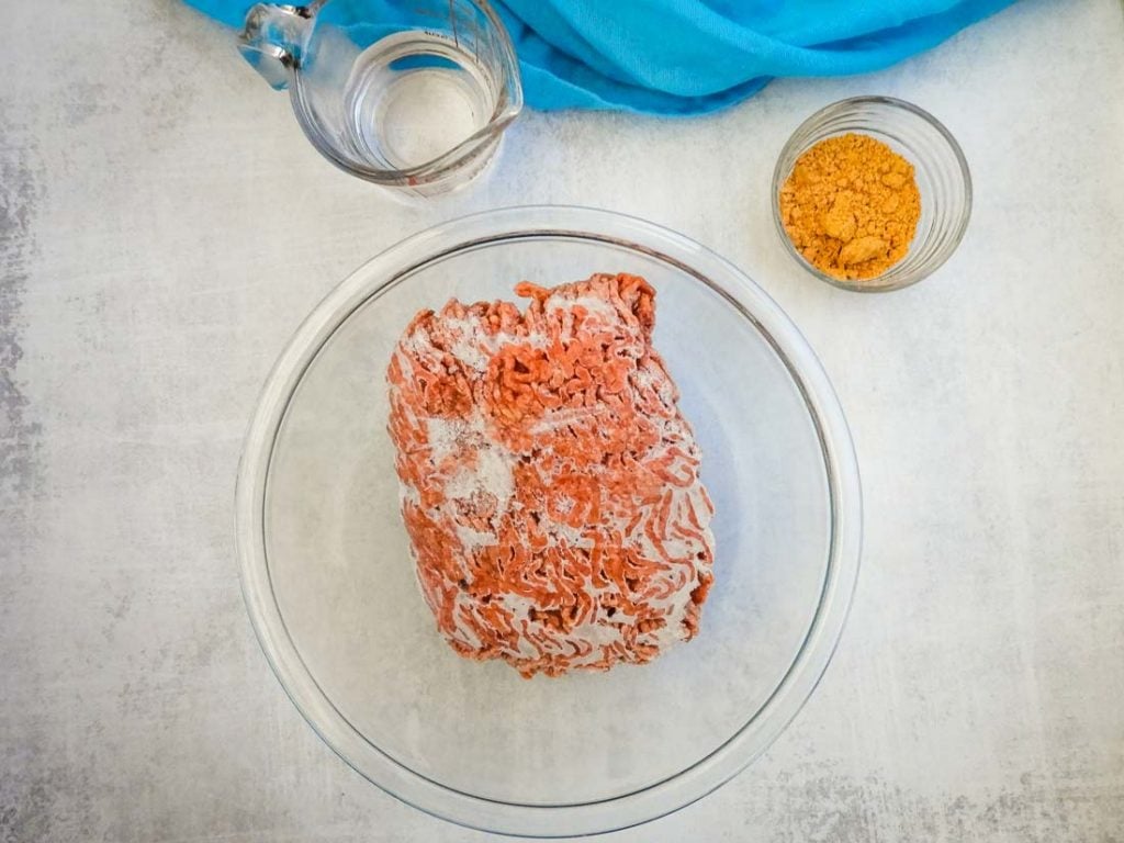 overhead photo of frozen ground beef, water, and seasoning in clear bowls with a blue linen