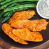 black plate with cooked boneless buffalo wings and green beans, with small white dish of ranch dressing on the side.