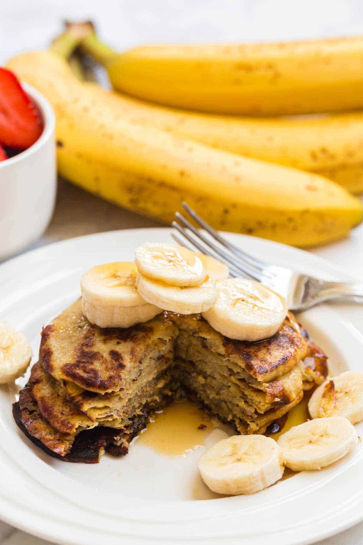 Stack of two ingredient pancakes on a white plate with sliced bananas and syrup on top. Whole bananas in the background.