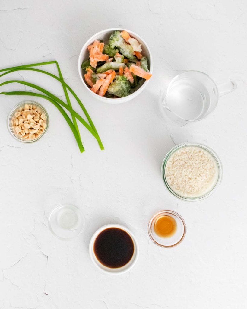Image of ingredients in fried rice recipe. Bowls of frozen vegetable, water, white rice, coconut aminos, and sesame oil. On the counter is a few stalks of green onions and a bowl of peanuts.