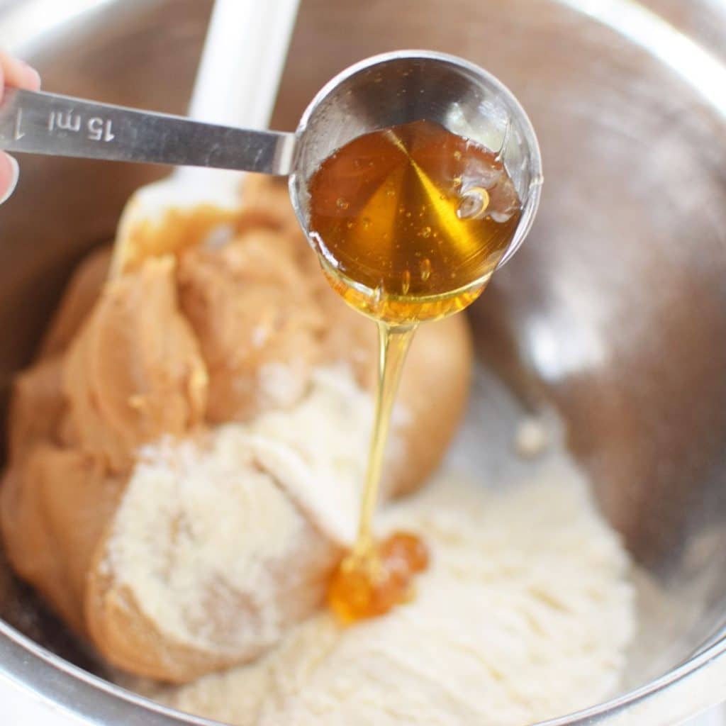 A stainless steel bowl with creamy peanut butter, coconut flour and a tablespoon of honey being added to it.