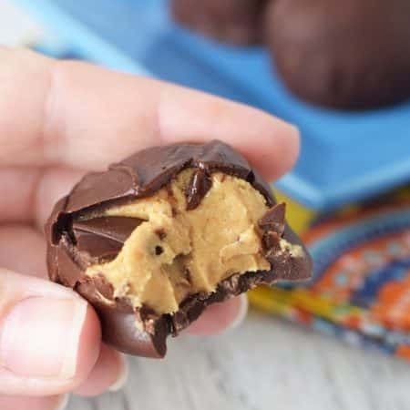 A hand holding a peanut butter buckeye that has a bite taken out of it. In the background there is a blue square plate with 5 more buckeyes.