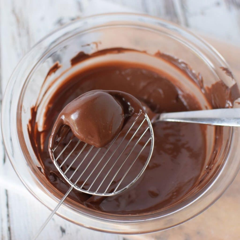 A glass bowl with melted chocolate inside it and a peanut butter ball that has just come out of the chocolate and is all covered with melted chocolate.