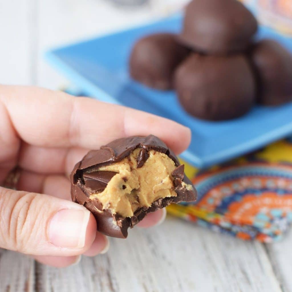 A hand holding a peanut butter buckeye that has a bite taken out of it. In the background there is a blue square plate with 5 more buckeyes.