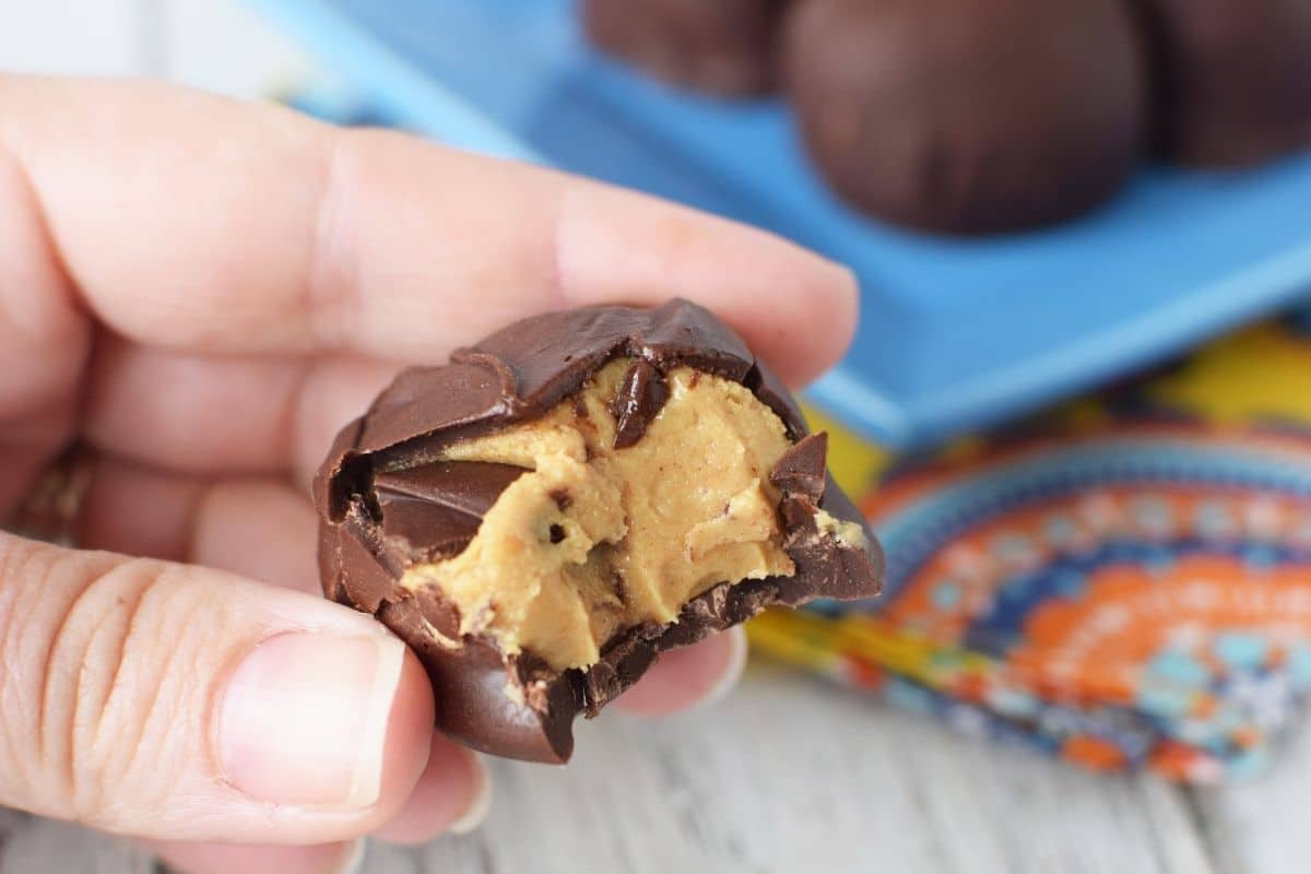 A hand holding a peanut butter buckeye that has a bite taken out of it. In the background there is a blue square plate with 5 more buckeyes.