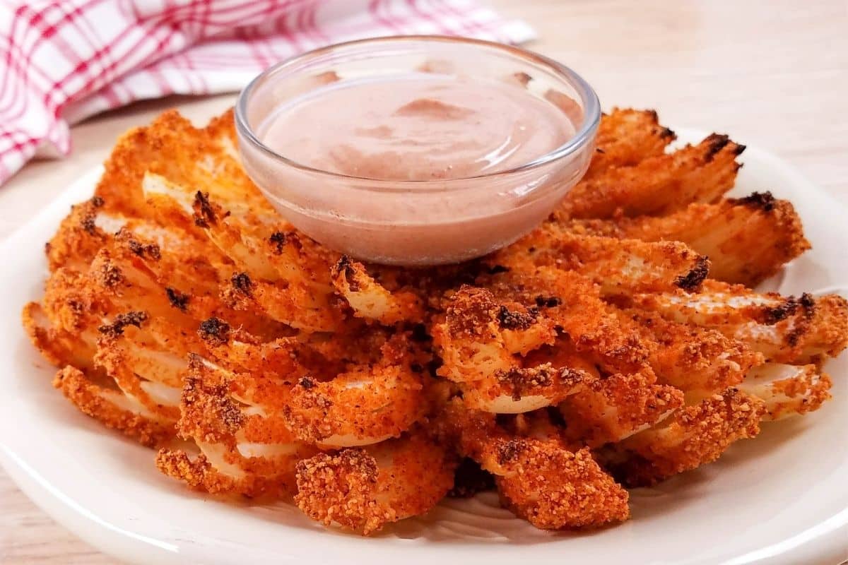 A white bowl with a cut and air fried blooming onion and in the center is a glass bowl of spicy dipping sauce.