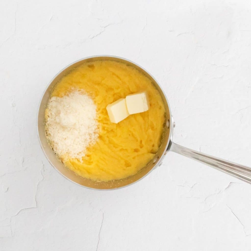 A white counter with a stainless steel saucepan that has cooked polenta, parmesan cheese, and butter inside.