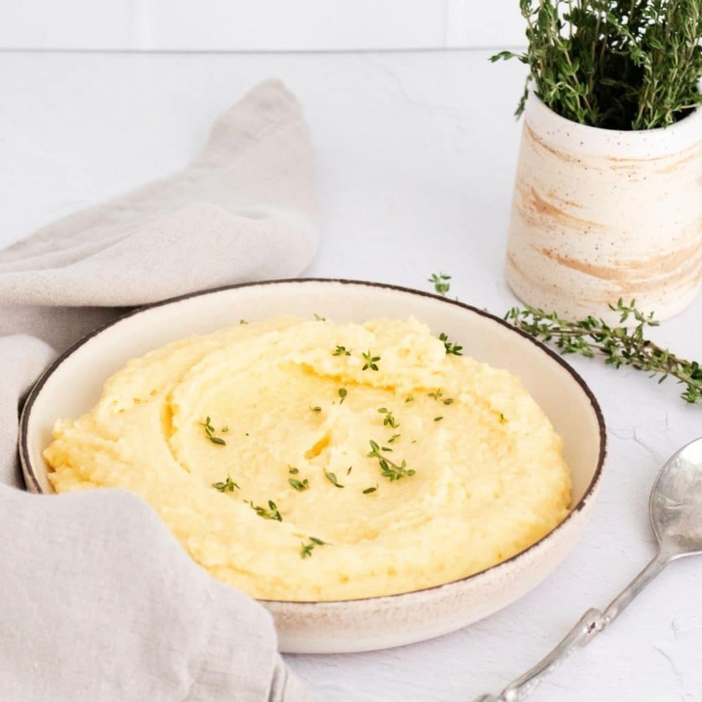 A neutral color counter and napkin with a bowl of creamy polenta with a sprinkle of thyme and off to the side is a cup of fresh thyme.