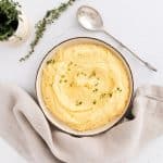 A neutral color counter and napkin with a bowl of creamy polenta with a sprinkle of thyme and off to the side is a cup of fresh thyme.