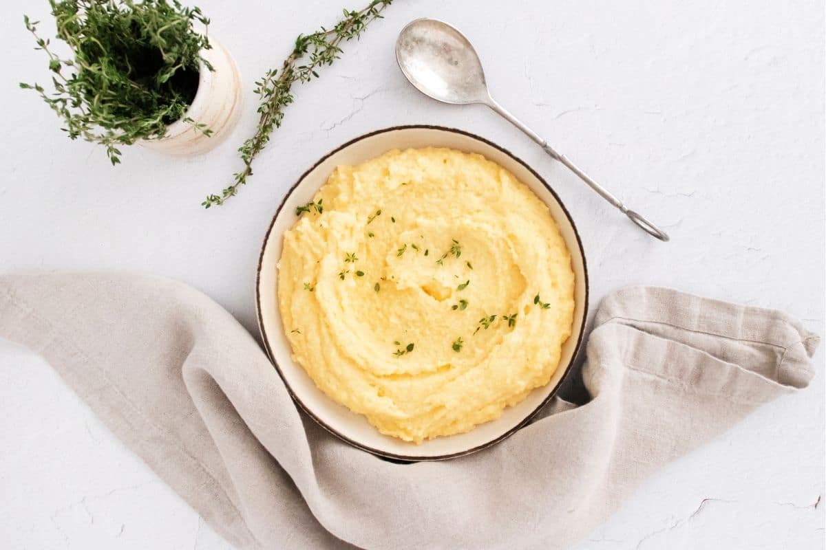 A neutral color counter and napkin with a bowl of creamy polenta with a sprinkle of thyme and off to the side is a cup of fresh thyme.
