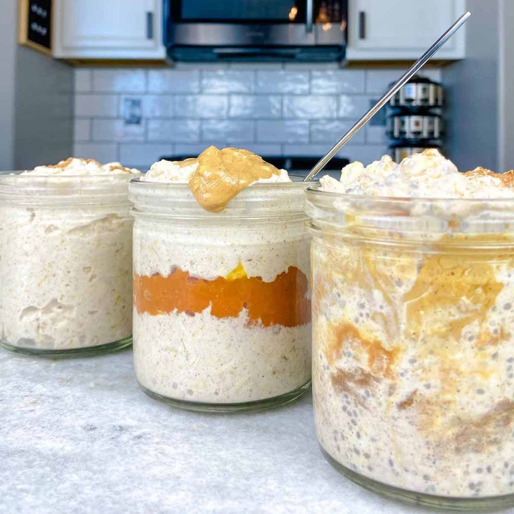 Several versions of pumpkin spice overnight oats on a countertop, including one with a pumpkin layer and one mixed with peanut butter.