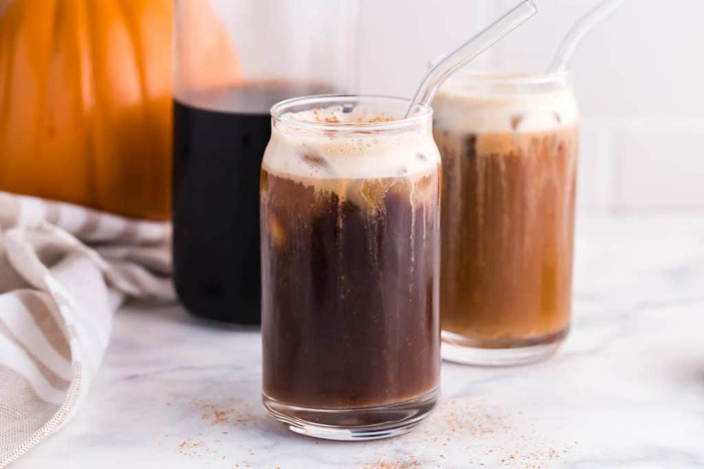carafe of cold brew with two clear glasses of pumpkin cream cold brew in front. real pumpkin in the back left of photo. photo taken on white marble countertops.