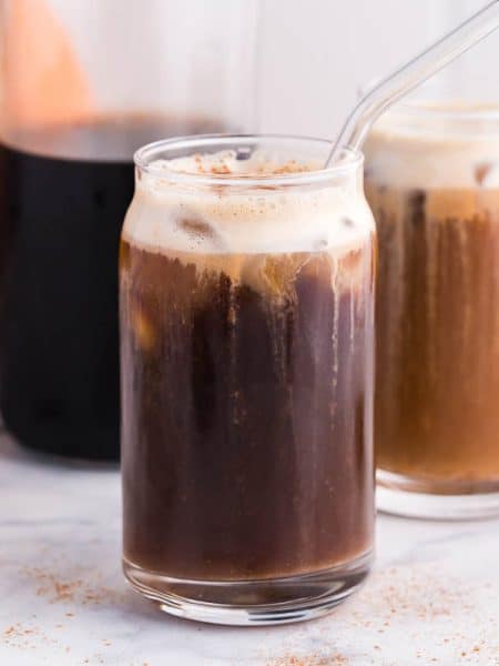 carafe of cold brew with two clear glasses of pumpkin cream cold brew in front. real pumpkin in the back left of photo. photo taken on white marble countertops.