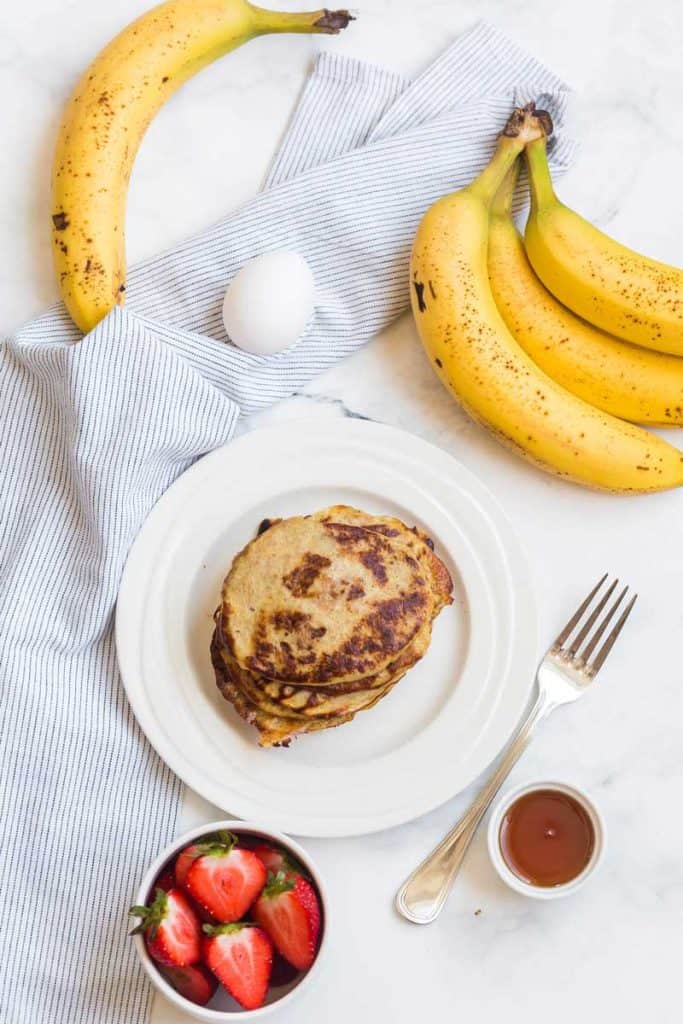 strawberries in a small bowl, stack of banana pancakes on a white plate, and whole bananas and eggs on a white background