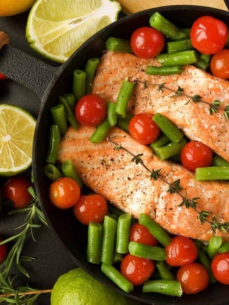 salmon and vegetables in a cast iron skillet, black countertop and sliced limes outside of pan