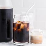 carafe of cold brew with a clear glass of cold brew with ice cubes. there is a small jar of cream on the right side of the pictures.