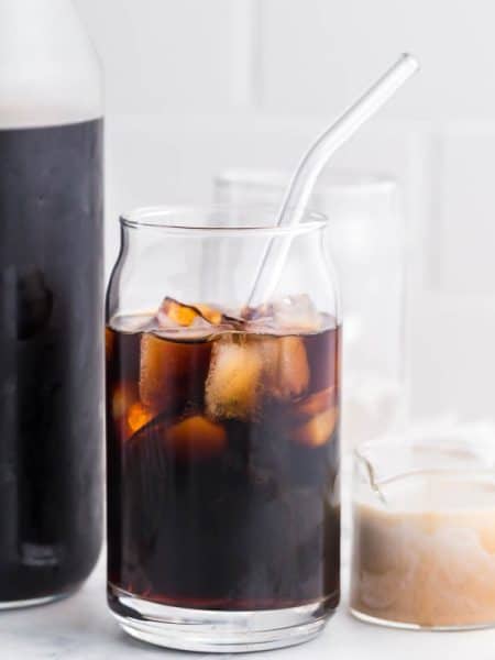 carafe of cold brew with a clear glass of cold brew with ice cubes. there is a small jar of cream on the right side of the pictures.