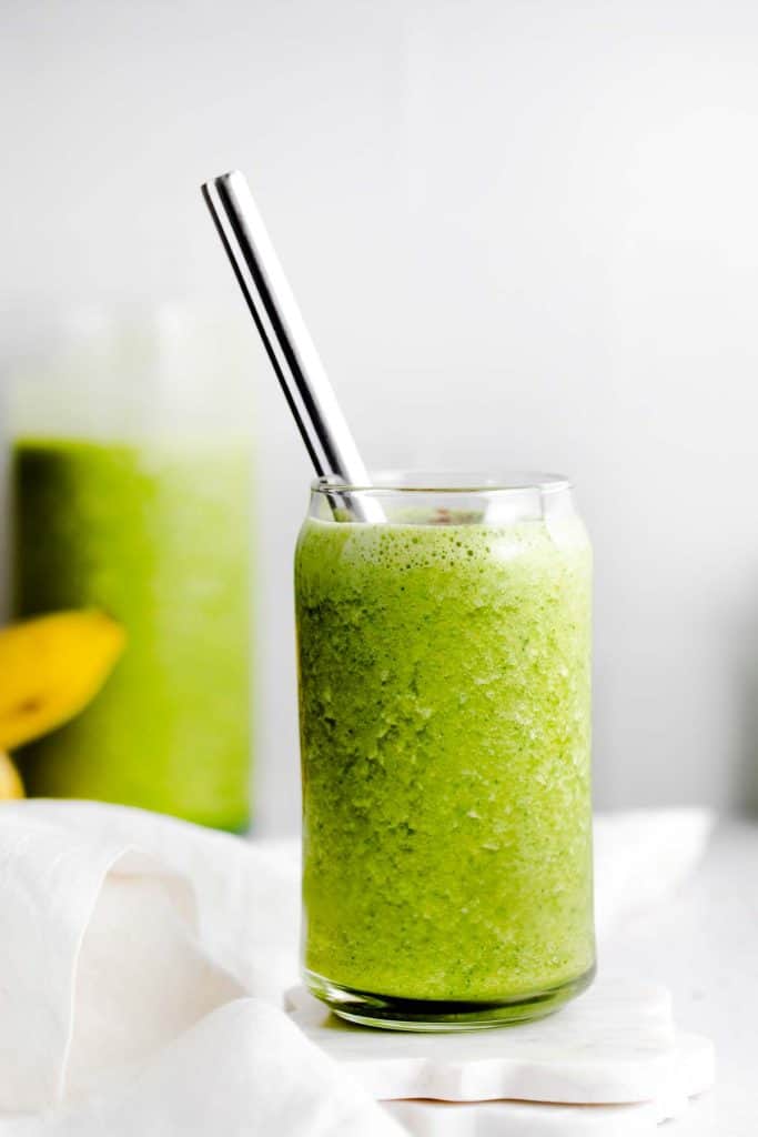 Glass full of green smoothie with a metal straw and bananas in the background. 