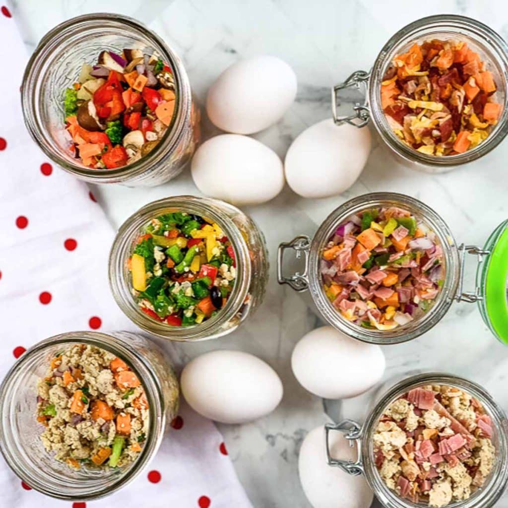 Six glass jars with ingredients for DIY Just Crack an Egg inside. Eggs are on the counter and a red polka dotted napkin is under the jars. 