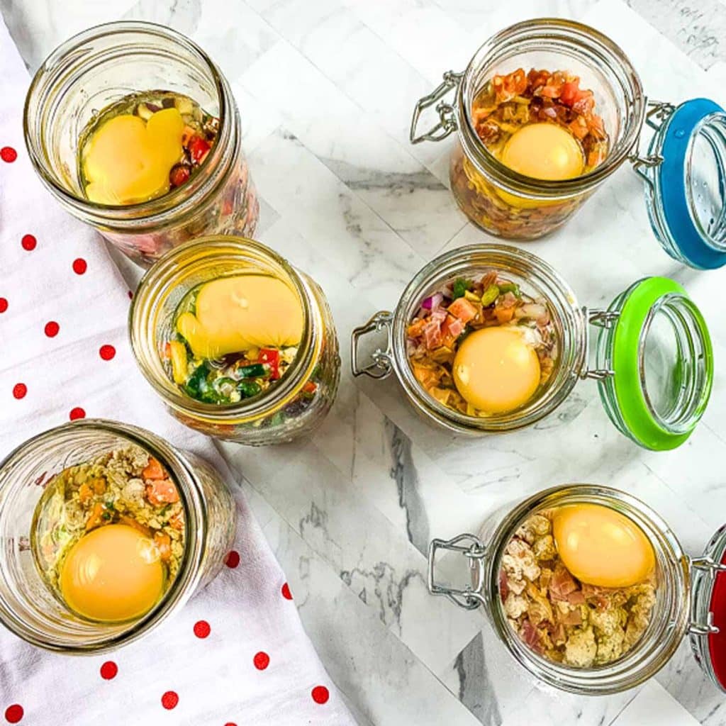 Six glass jars with ingredients for mini egg scrambles and eggs inside. 