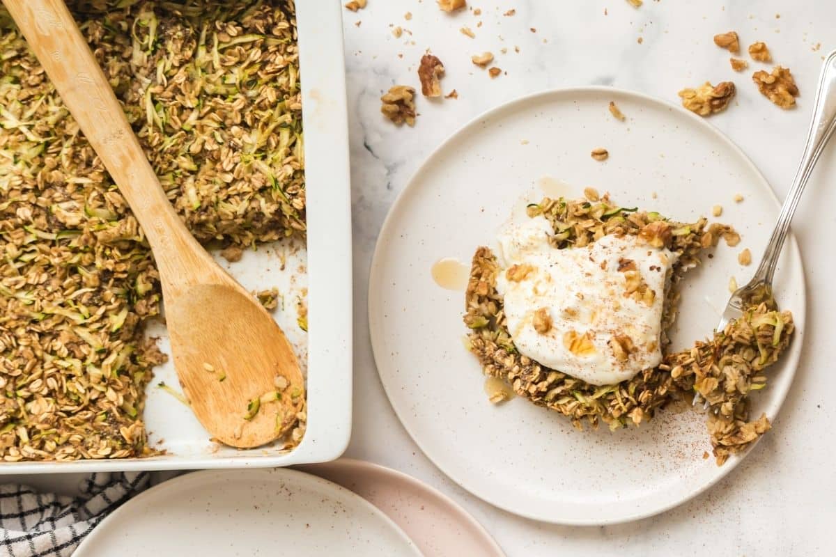 A white baking dish with a piece of zucchini bread baked oatmeal taken out of it with a wooden spoon and set on a circular white plate and then topped with whipped topping.
