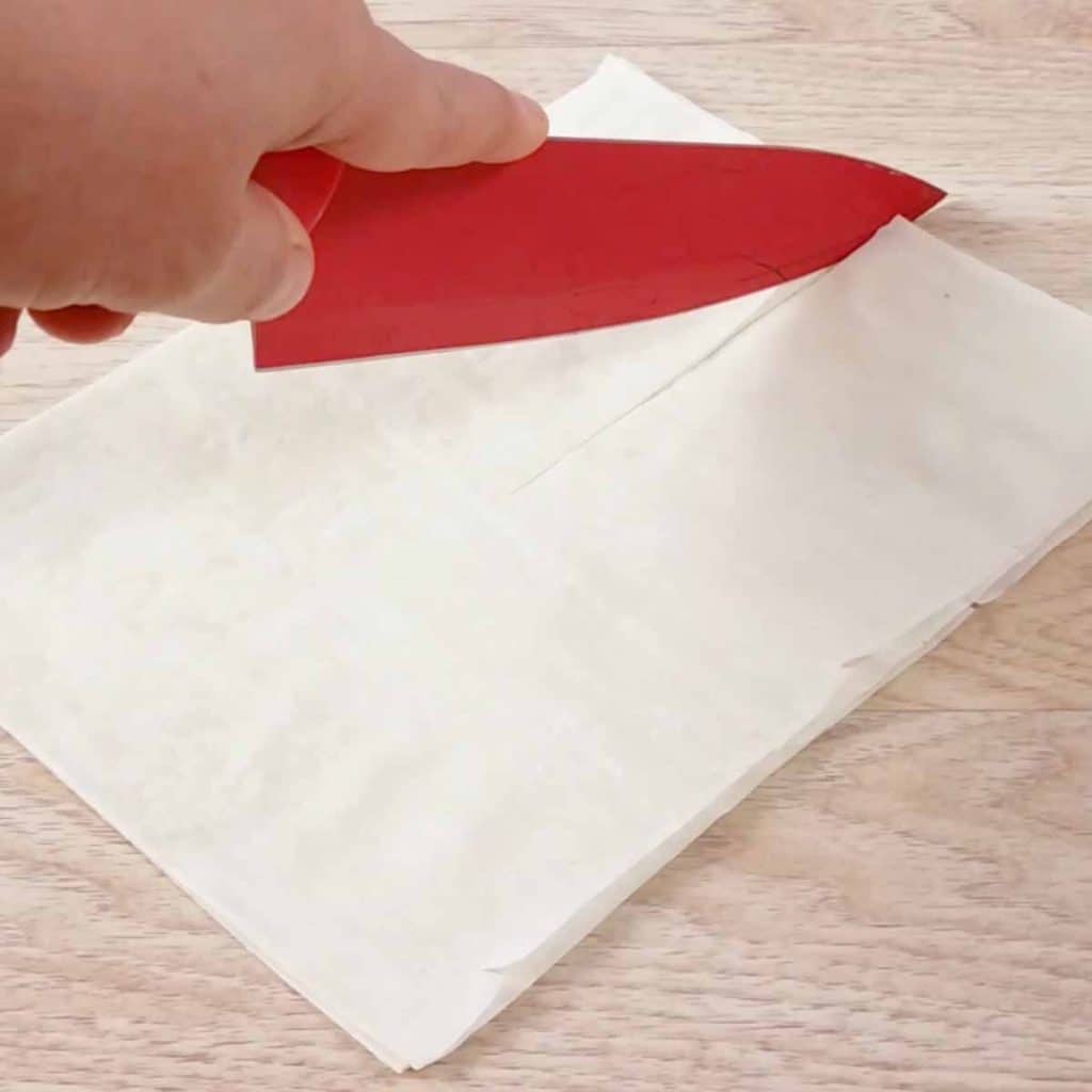 Hand cutting phyllo dough with a red plastic knife on a wood countertop. 