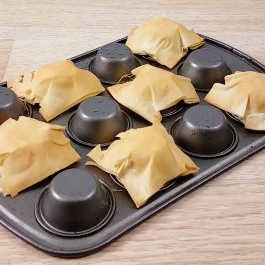 Upside down muffin tin with cooked phyllo dough cups pressed around every other cup. 