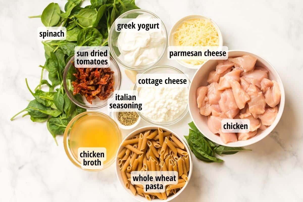 White counter with a scattering of spinach leaves along with bowls full of labeled ingredients needed to make tuscan chicken pasta recipe.