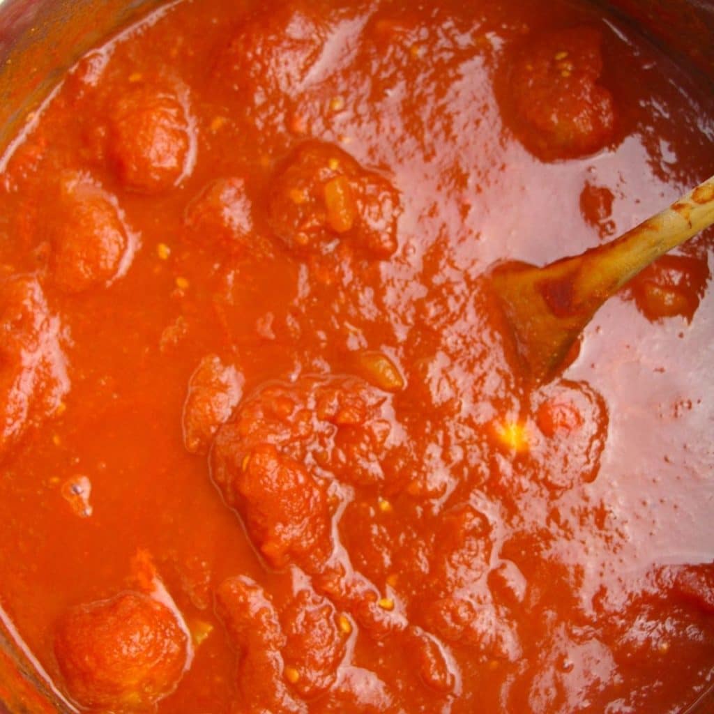 Homemade spaghetti sauce being stirred by a wooden spoon inside a stainless steel saucepan.