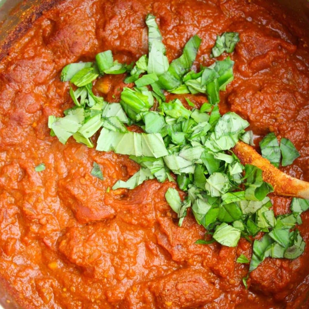 A stainless steel saucepan with spaghetti sauce and chopped fresh basil being stirred in with a wooden spoon.