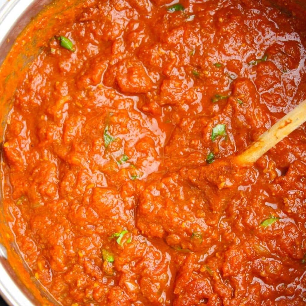 Homemade spaghetti sauce being stirred by a wooden spoon inside a stainless steel saucepan.