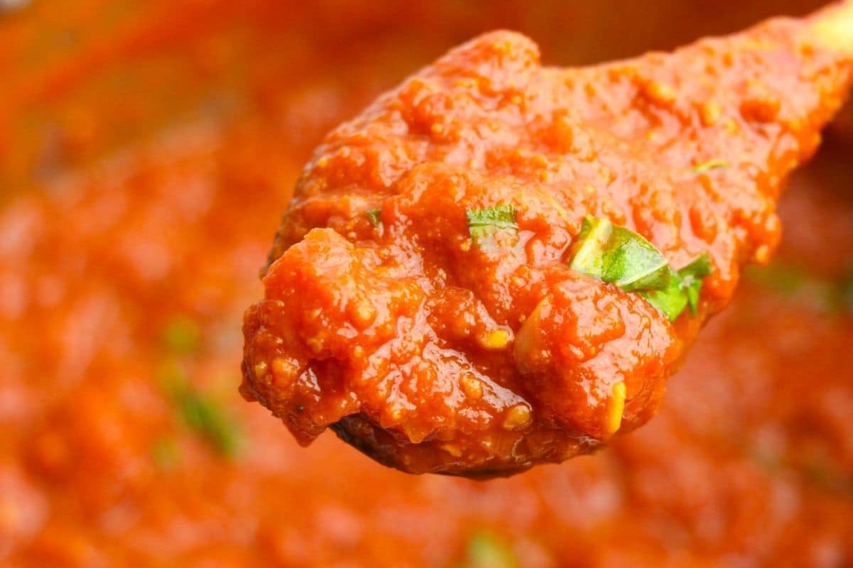 A spoon with spaghetti sauce and chopped fresh basil. There is a pot of sauce in the background.