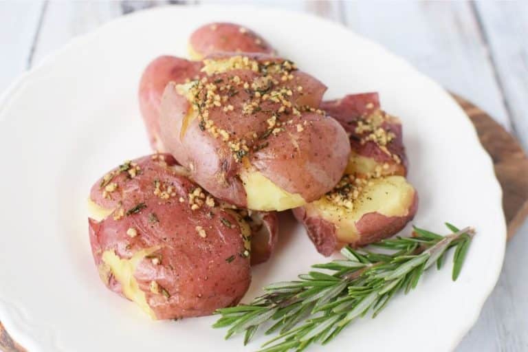 A white plate with three baked and smashed red potatoes that are covered in garlic and have a sprig of rosemary on the plate.