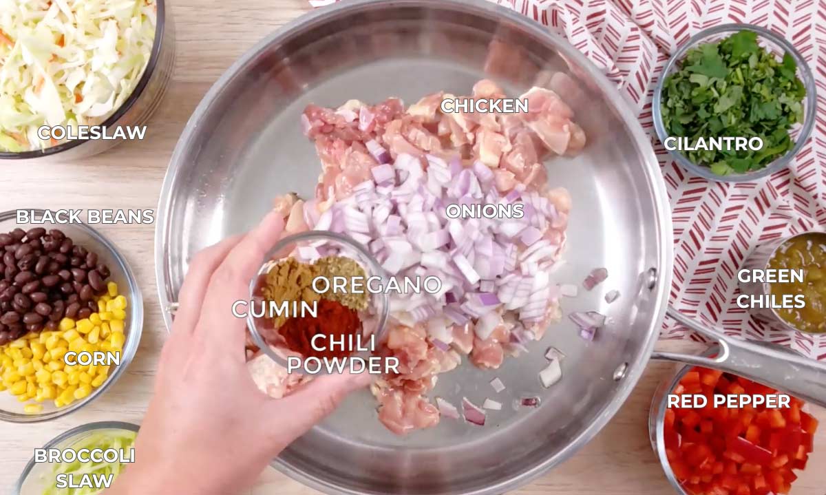 Steel pan with raw diced chicken and diced red onion. Spices, coleslaw, black beans and corn, cabbage, diced cilantro, green chiles, and red pepper are in clear bowls.