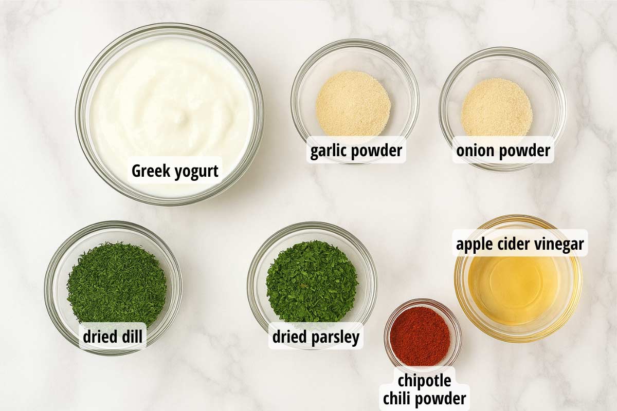 A marble countertop with small clear bowls full of ingredients: greek yogurt, garlic powder, onion powder, dried dill and parsley, and chipotle chili powder. 