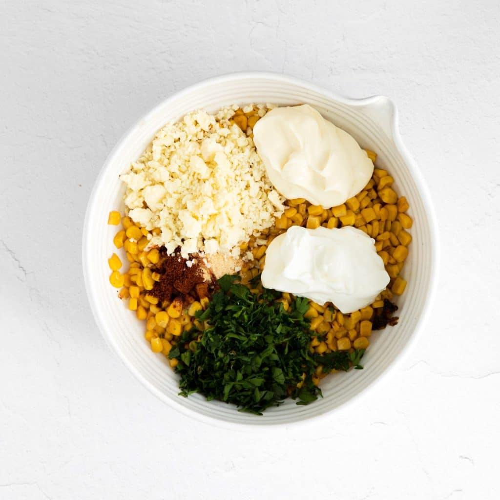 A white bowl full of corn and then topped with sections of cotija cheese, greek yogurt, cottage cheese, parsley and spices.