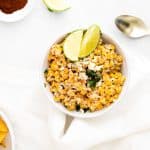 A white counter with a white bowl full of easy and healthy elote dip and topped with lime wedges and parsley. Off the the side is a spoon, a bowl of tortilla chips, another wedge of lime, and a bowl of spices.