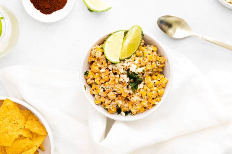 A white counter with a white bowl full of easy and healthy elote dip and topped with lime wedges and parsley. Off the the side is a spoon, a bowl of tortilla chips, another wedge of lime, and a bowl of spices.