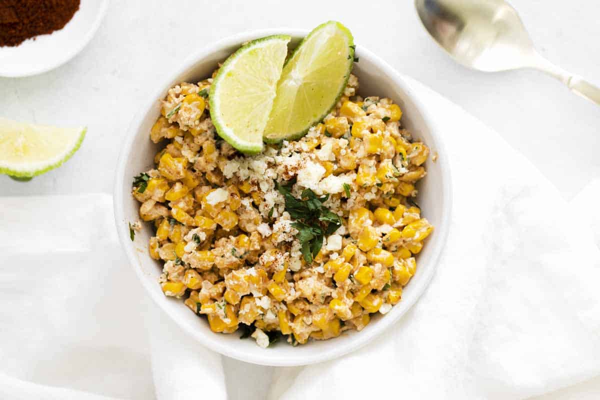 White background with white bowl of elote dip garnished with sliced limes.