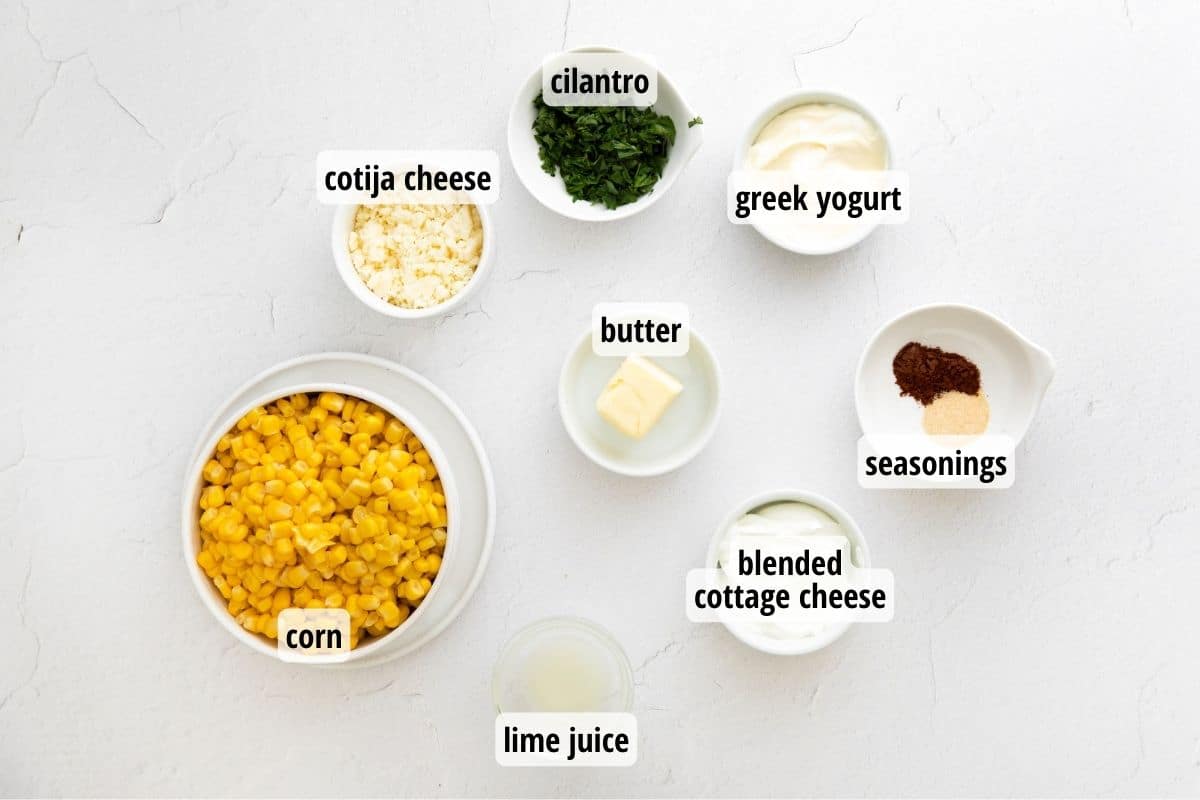 A white counter topped with bowls full of labeled ingredients including corn, cotija cheese, cilantro, greek yogurt, spices, cottage cheese, butter and lime juice.